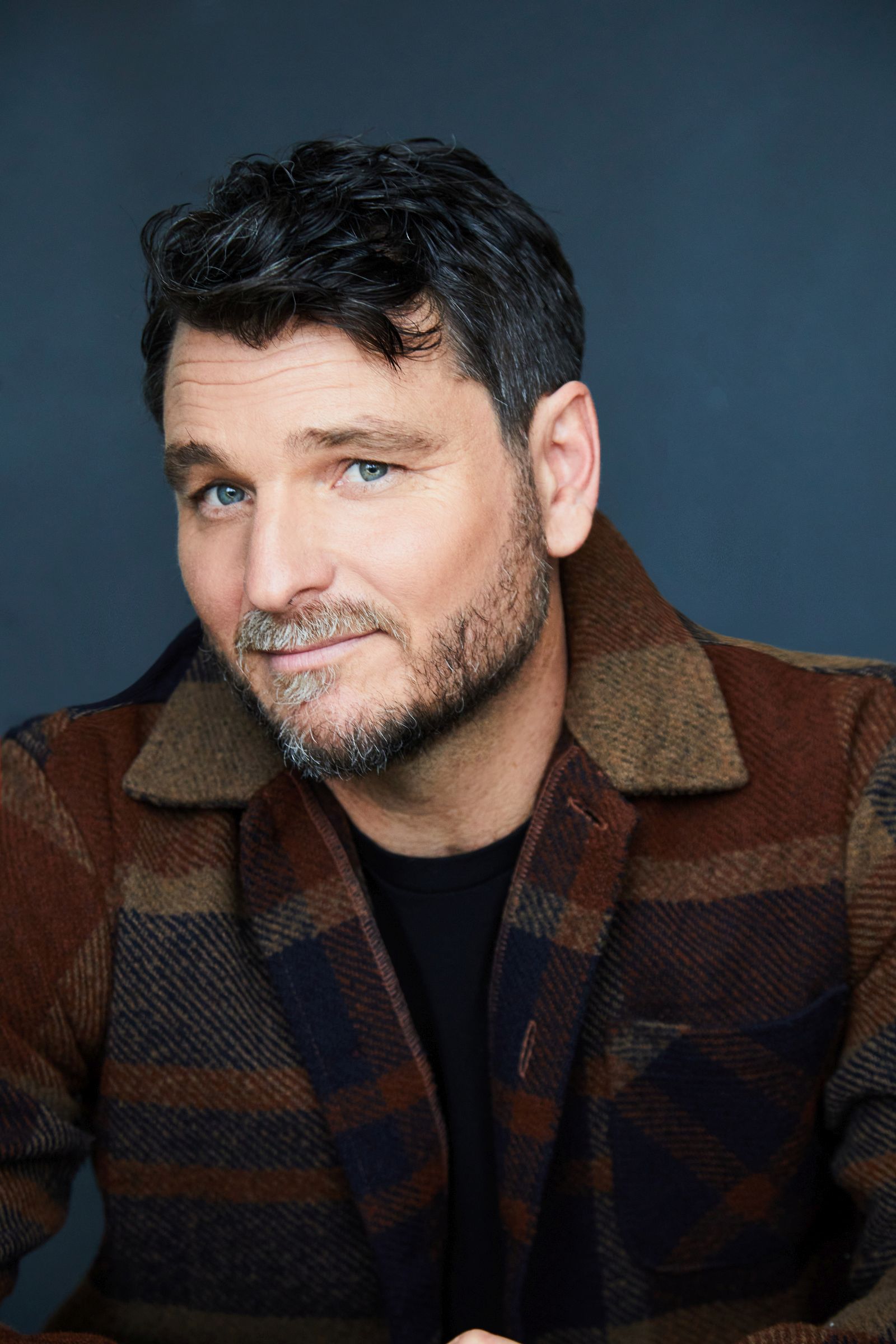 Man with short brown hair, wearing a plaid jacket, smiling slightly.