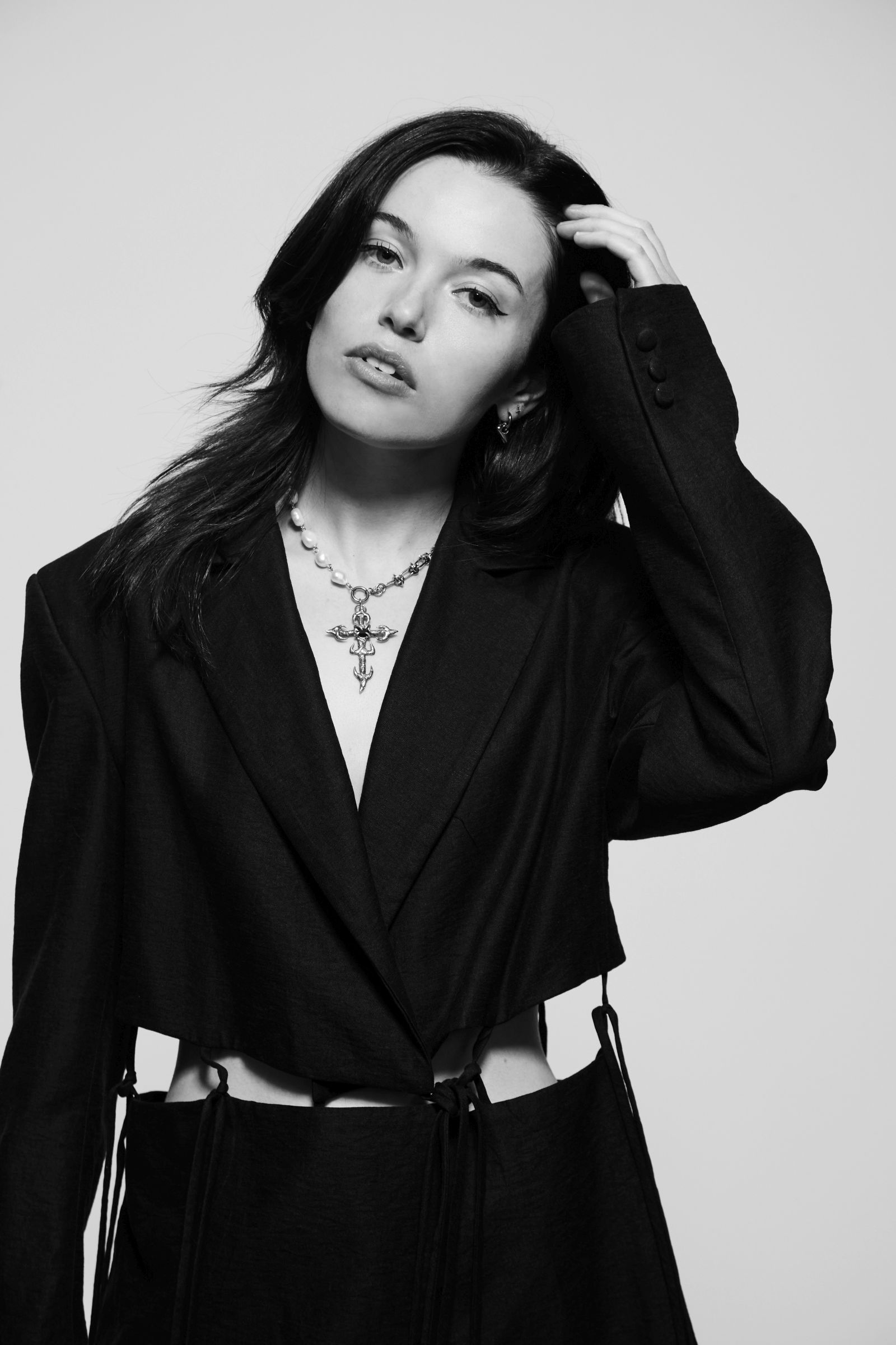 Woman in a black jacket with a pearl necklace and pendant, hand through hair.