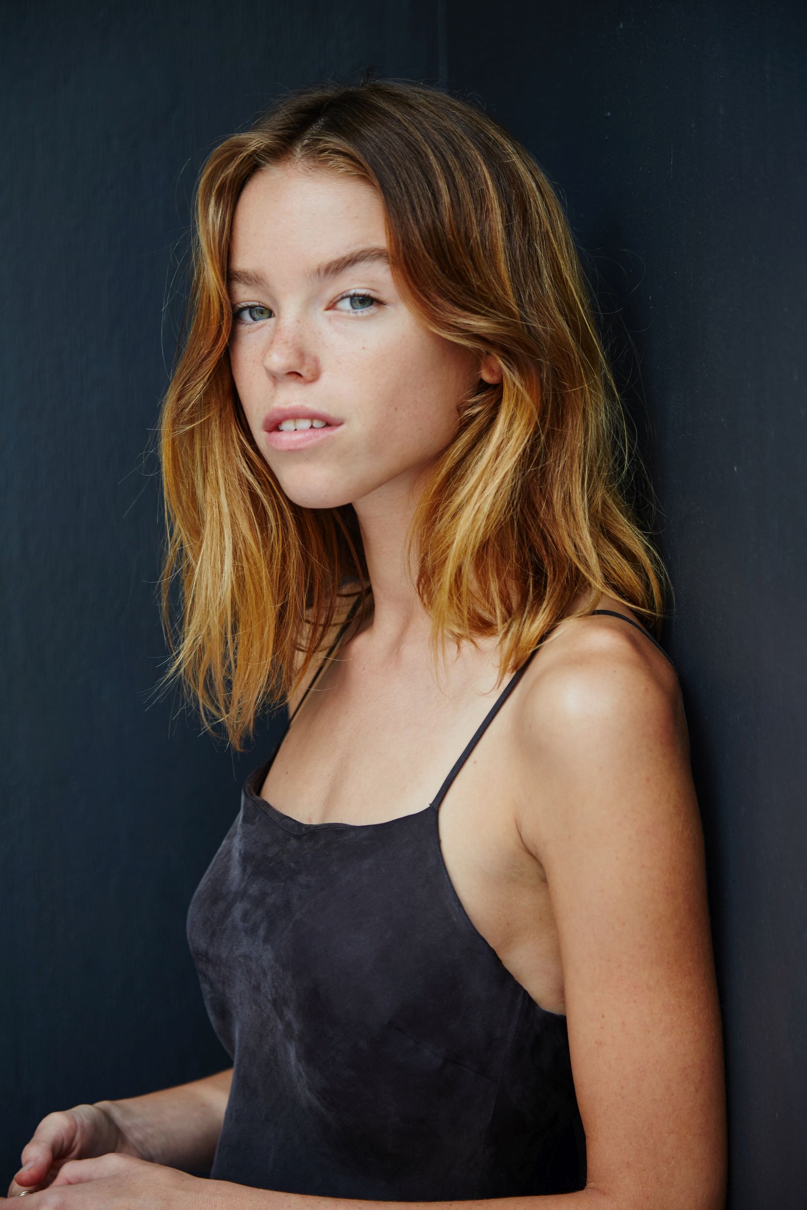 Woman with light brown hair in dark sleeveless dress against dark background.