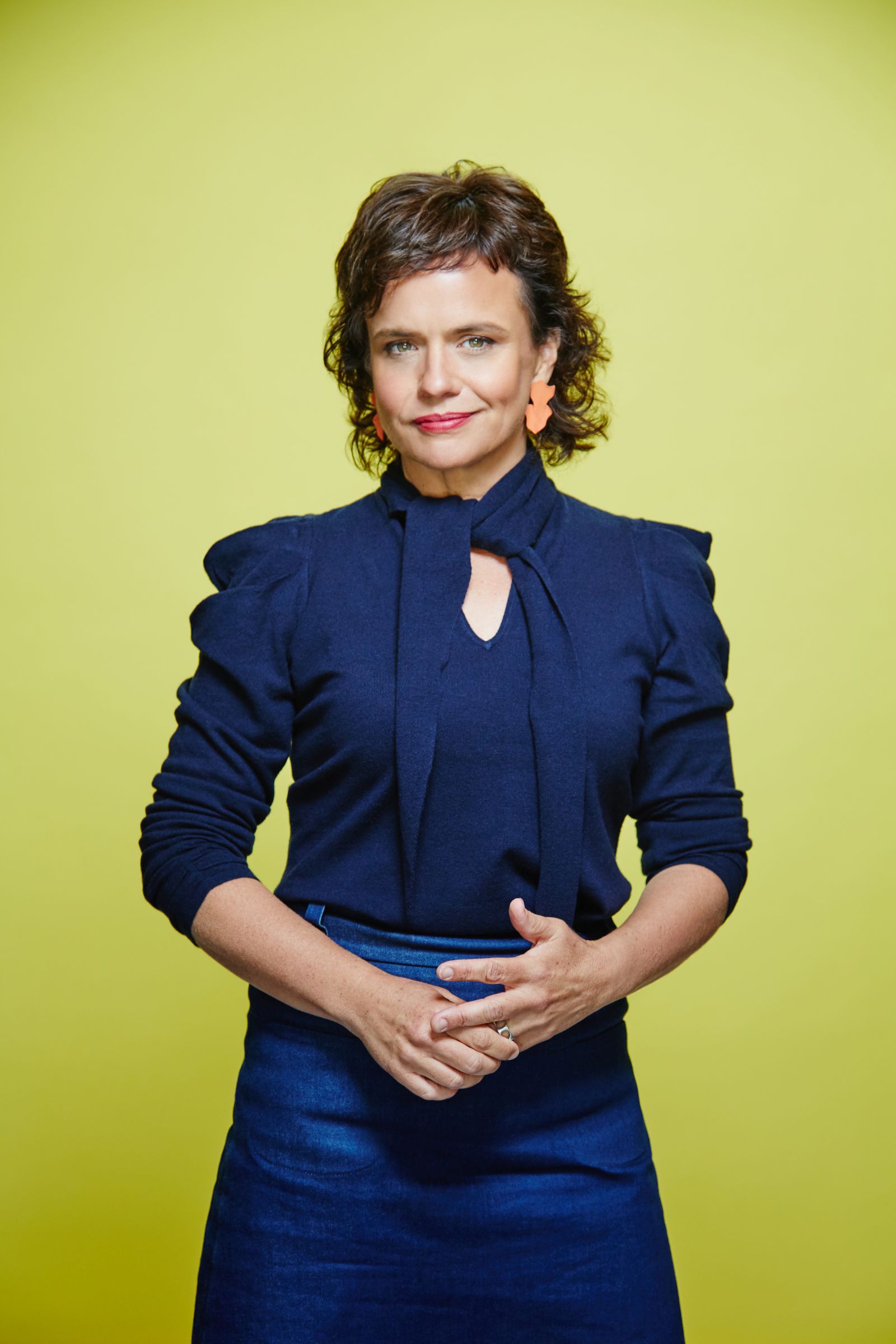 Mid-length studio portrait of person in navy top, orange earrings, yellow backdrop.