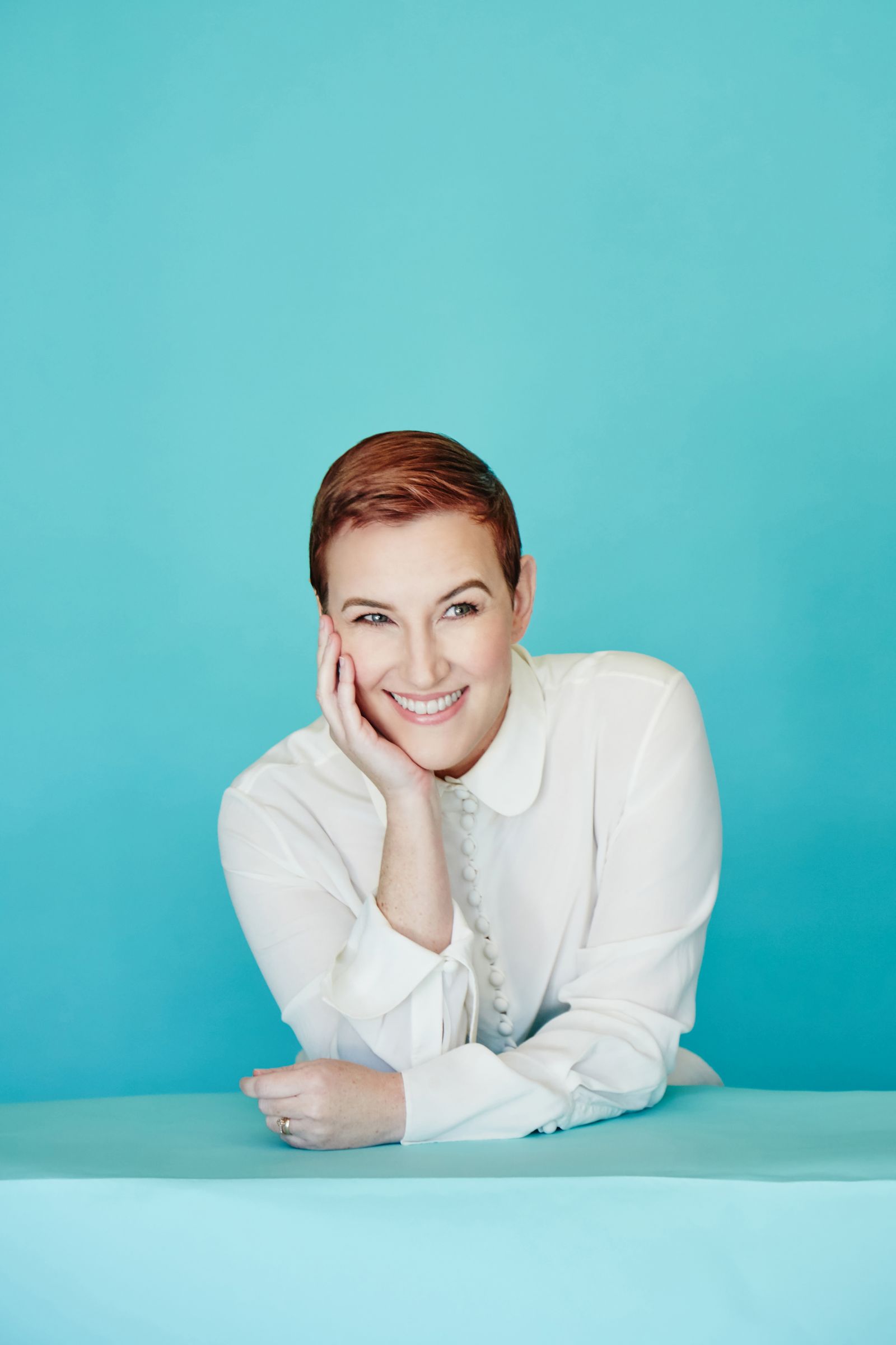 Smiling person with short hair in white shirt, leaning on hand, teal studio backdrop.