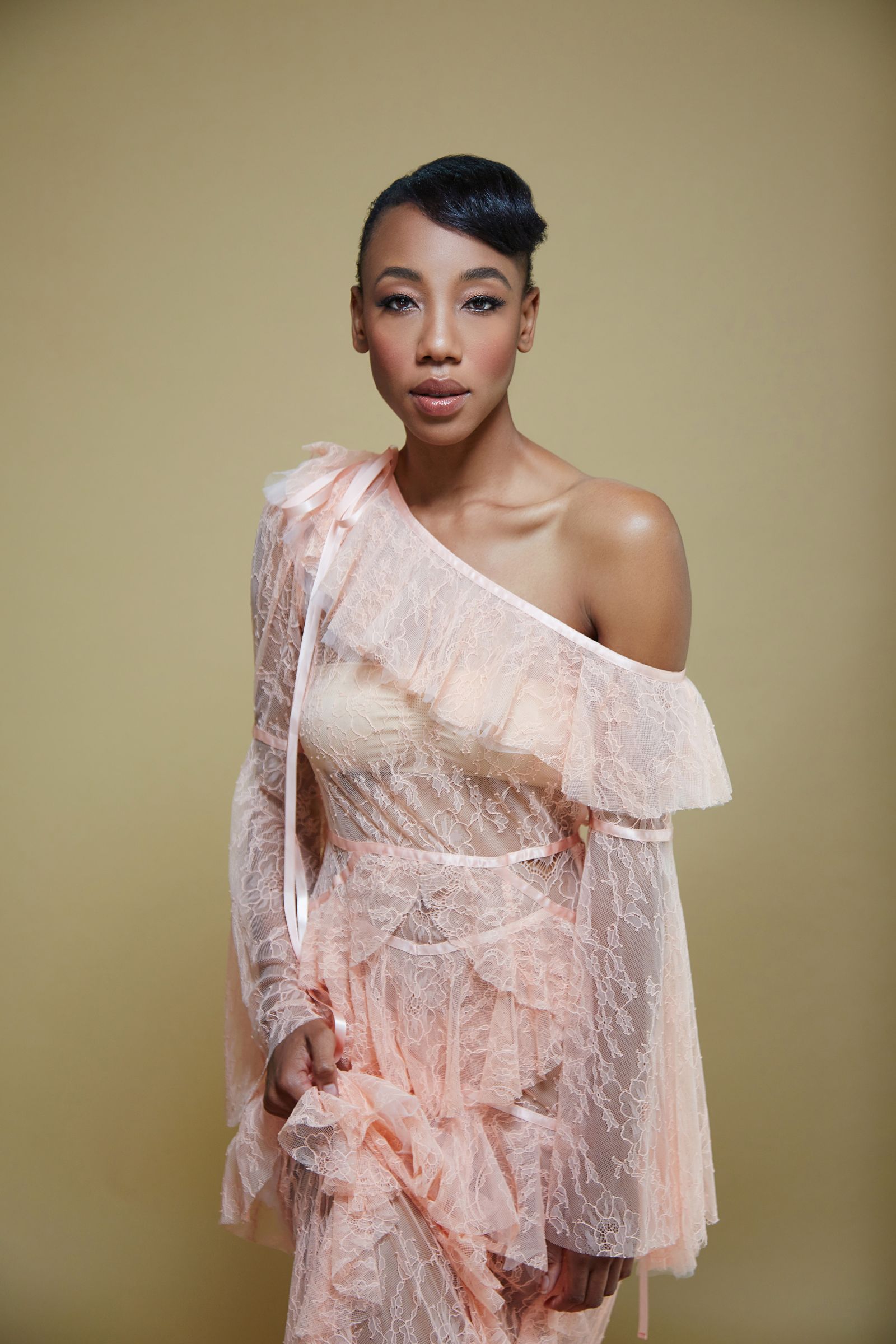 Studio portrait of model in peach sheer lace one-shoulder dress against beige backdrop.