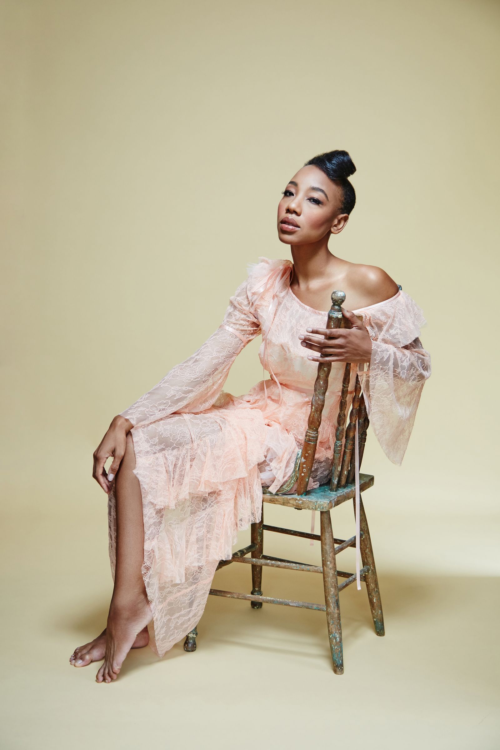 Seated woman in off-shoulder peach lace gown on rustic chair, barefoot, studio backdrop.