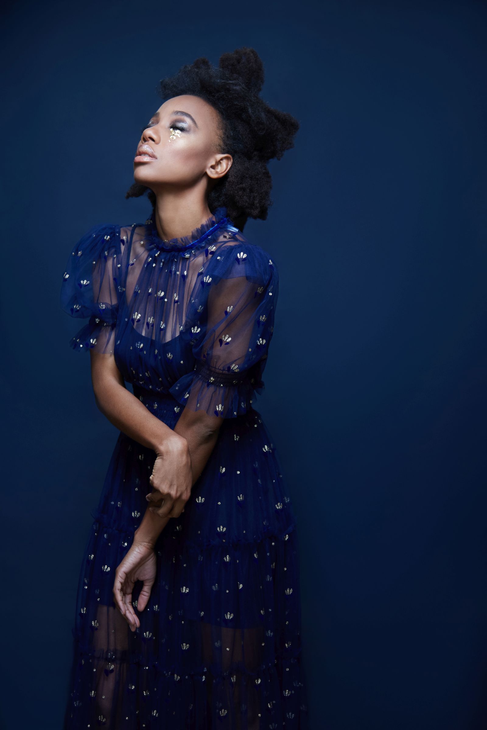 Studio editorial portrait of person in sheer navy gown, glitter tear, upward gaze, deep blue backdrop.