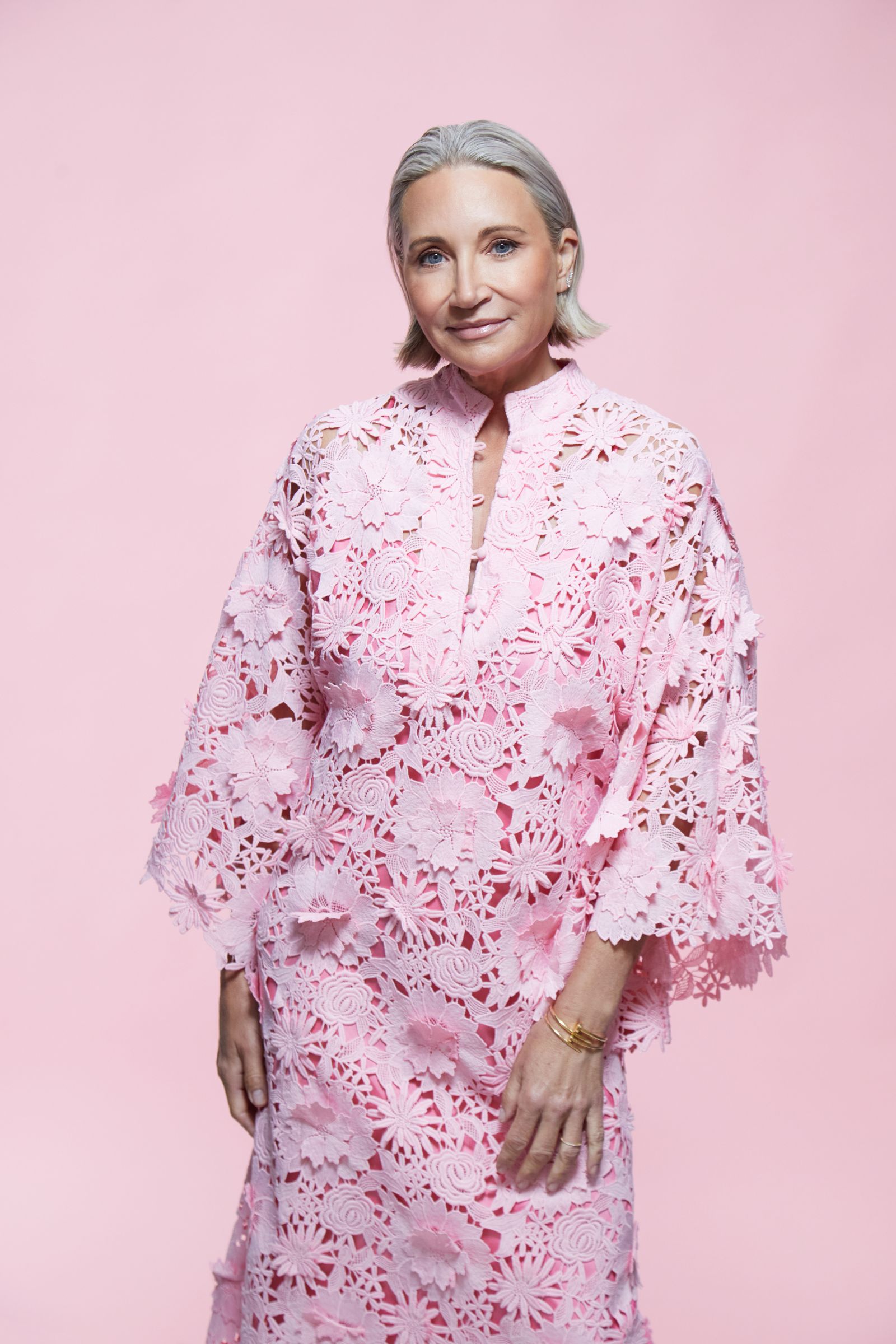 Three-quarter studio portrait of woman with short silver hair in pink floral lace dress on pink backdrop.