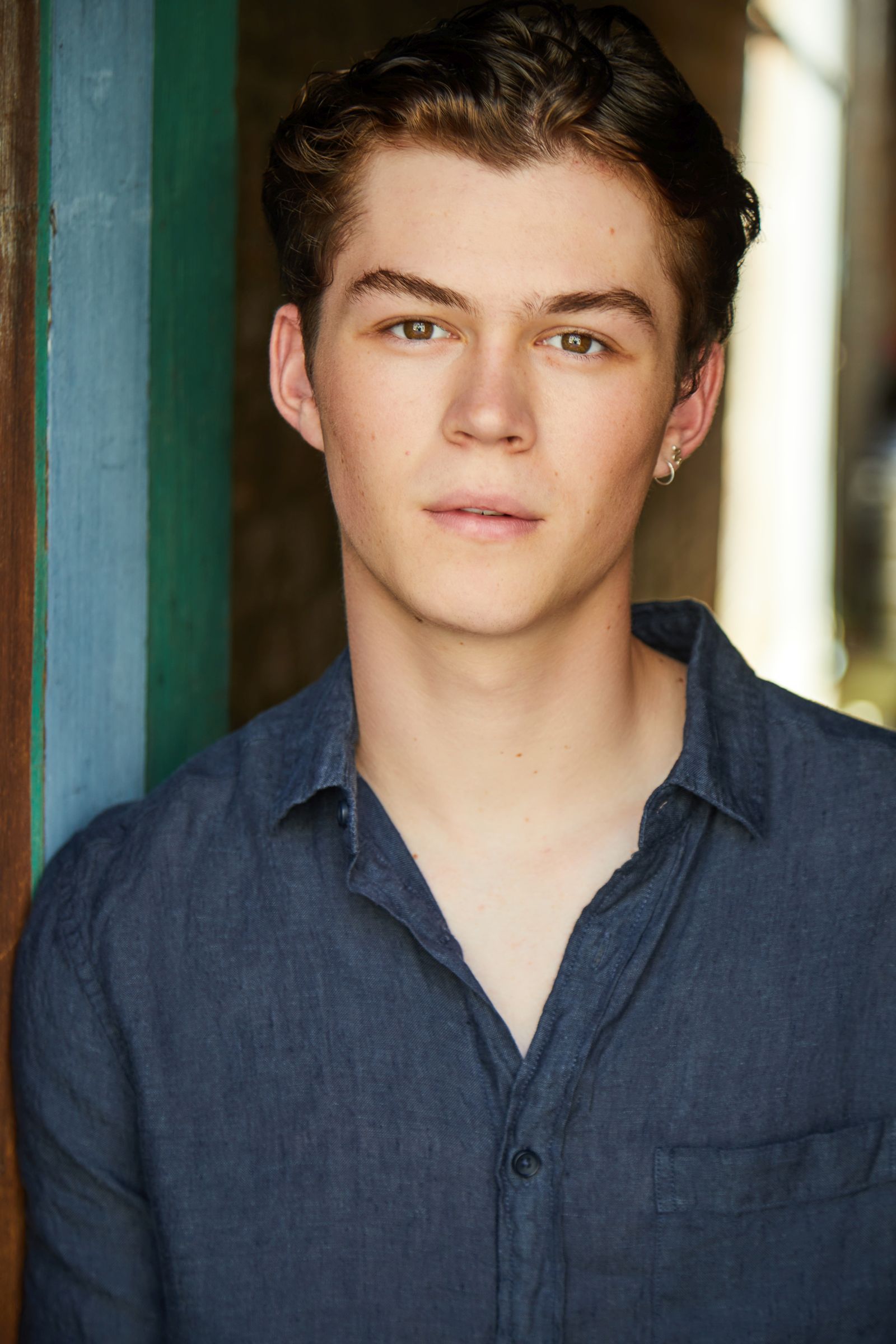 Young person with short hair and earring wearing a blue shirt, photographed indoors.