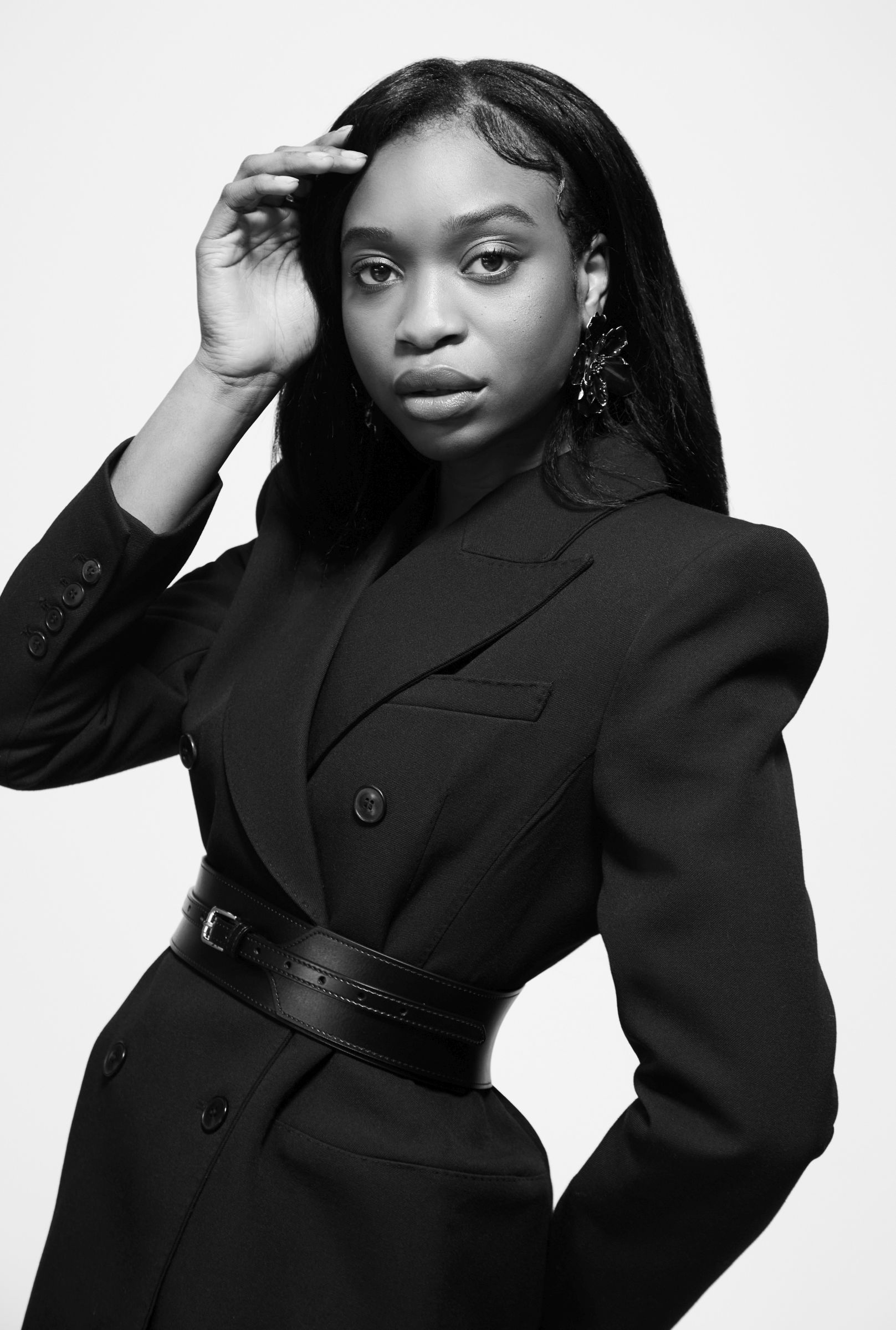 Black-and-white editorial studio portrait of person in belted blazer and statement earrings.