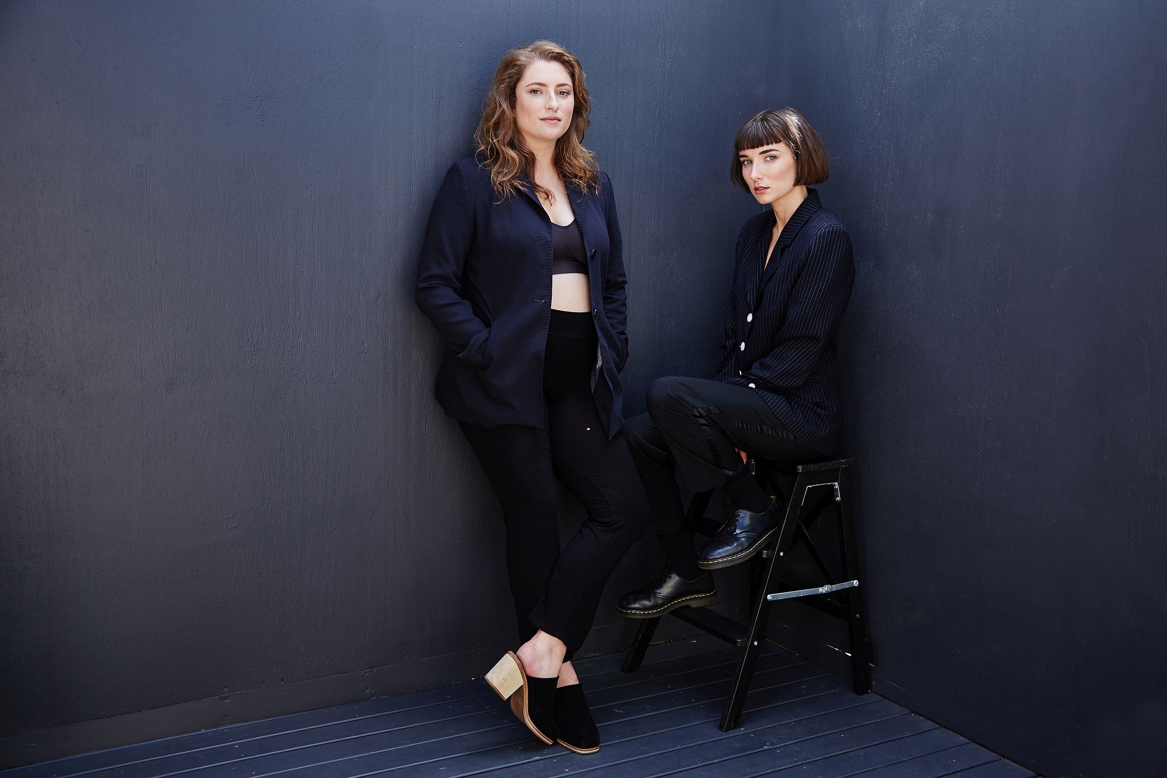 Editorial-style portrait of two people in black; one standing, one seated on a stool against a dark wall.