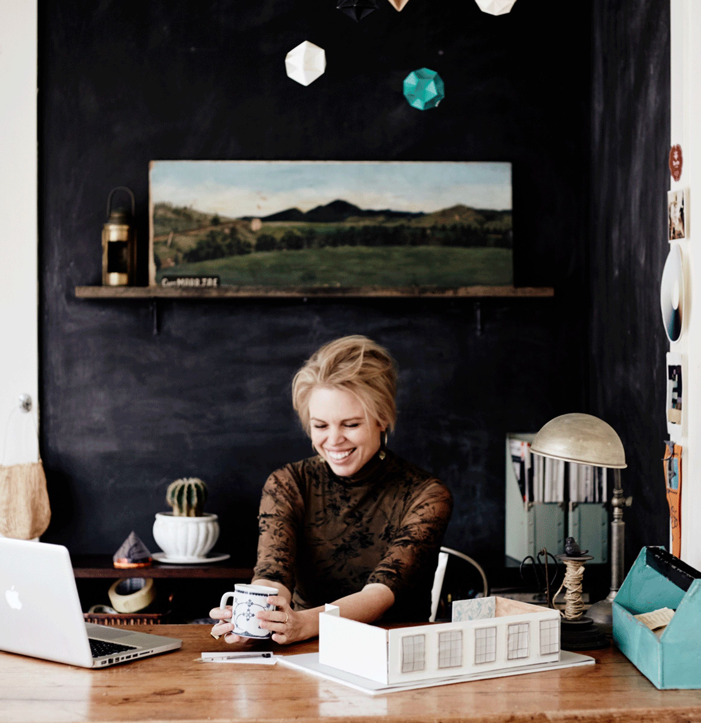 Smiling creative at studio desk with laptop, coffee mug, and architectural model.