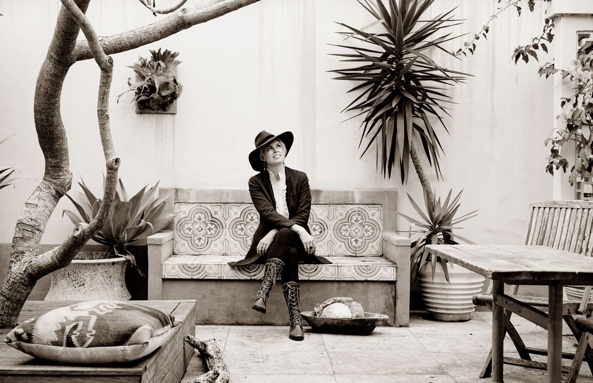 Sepia editorial portrait: person in wide-brim hat, cross-legged on tiled bench, courtyard plants.